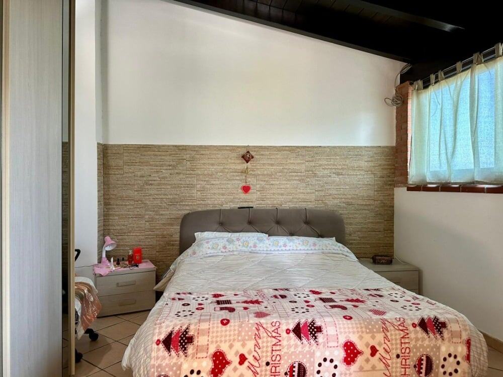Spazio notte matrimoniale con letto confortevole, decorazioni natalizie, rivestimento in pietra sulla parete e finestra con tende leggere, ambiente accogliente e semplice.