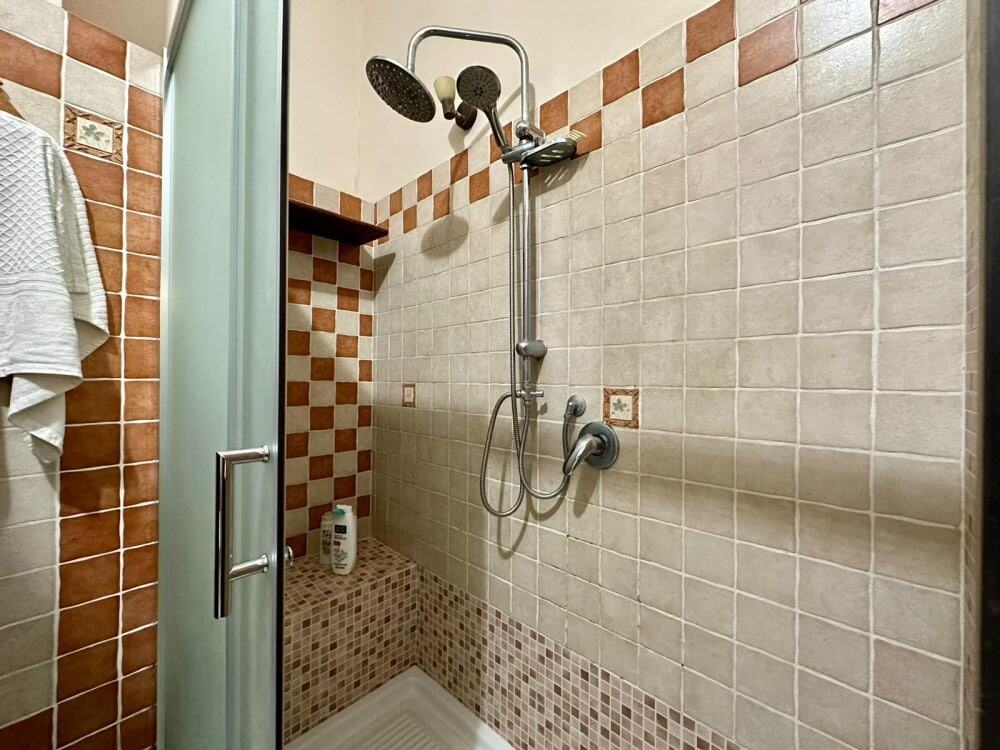 Doccia con pioggia a soffitto in bagno, rivestimento in piastrelle beige e terracotta, spazio doccia angolare con accessori moderni, ideale per ristrutturazioni e arredo bagno.