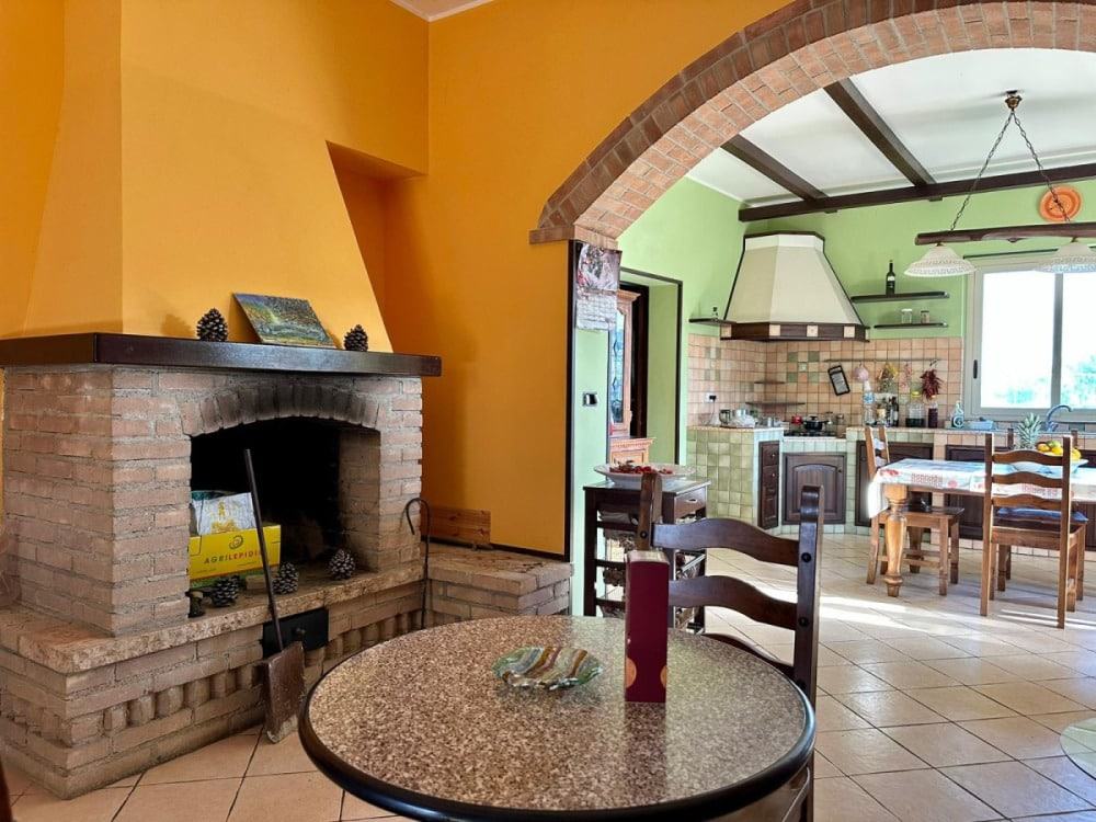 Accogliente cucina in stile rustico con arredi in legno, pareti verdi e gialle, camino in mattoni, grande finestra per la luce naturale, atmosfera calda e familiare.