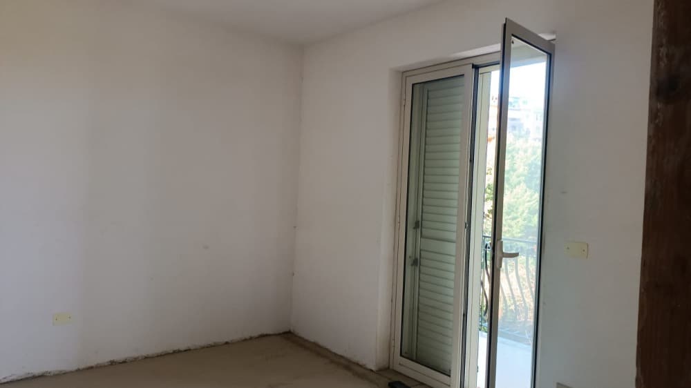 Vano vuoto con porta finestra che dà su balcone, pareti bianche e pavimento grezzo, spazio ideale per ristrutturazione e arredo, luminoso ambiente abitativo da personalizzare.