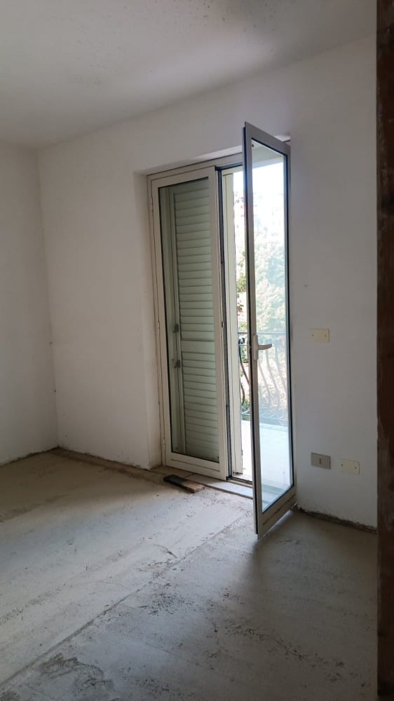 Completo interno di una stanza in fase di ristrutturazione con porta finestra a battente che si apre su un balcone con vista panoramica, pareti bianche e pavimento grezzo in cemento.