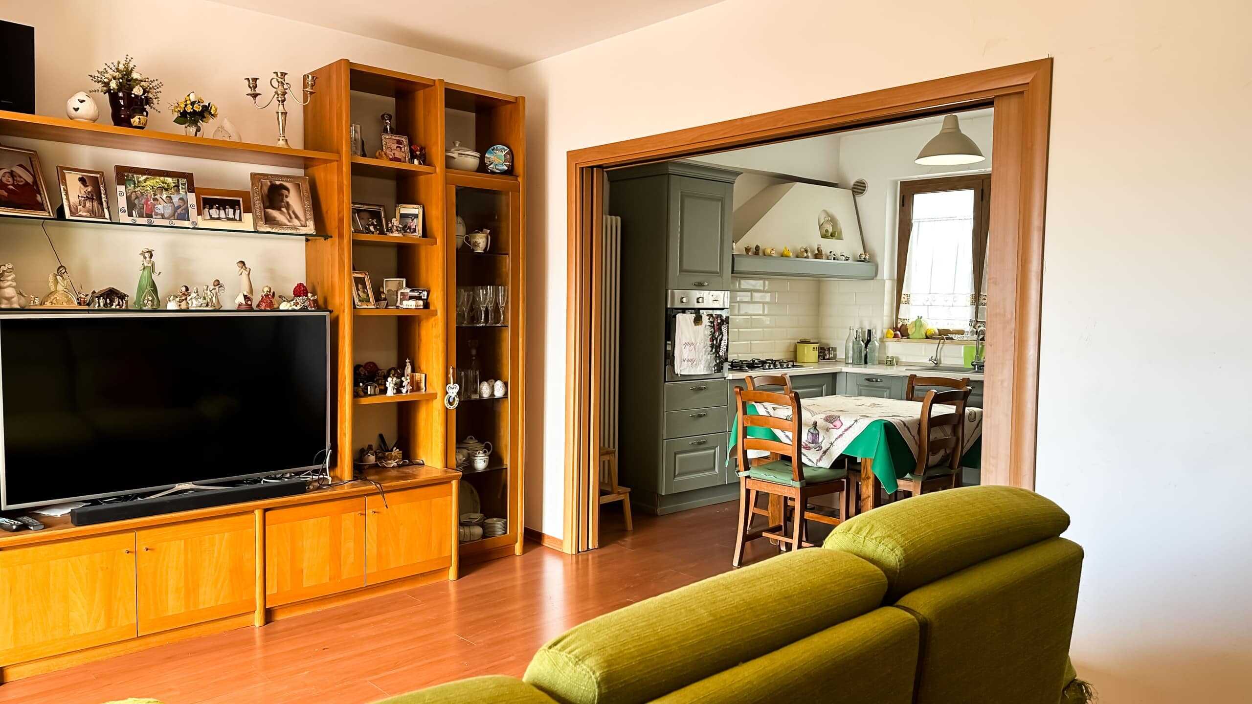 Classe di merito: divano in salotto con mobiletto e scaffali pieni di decorazioni e fotografie, vista sulla cucina moderna con mobili verdi e tavolo apparecchiato, atmosfera calda e accogliente.