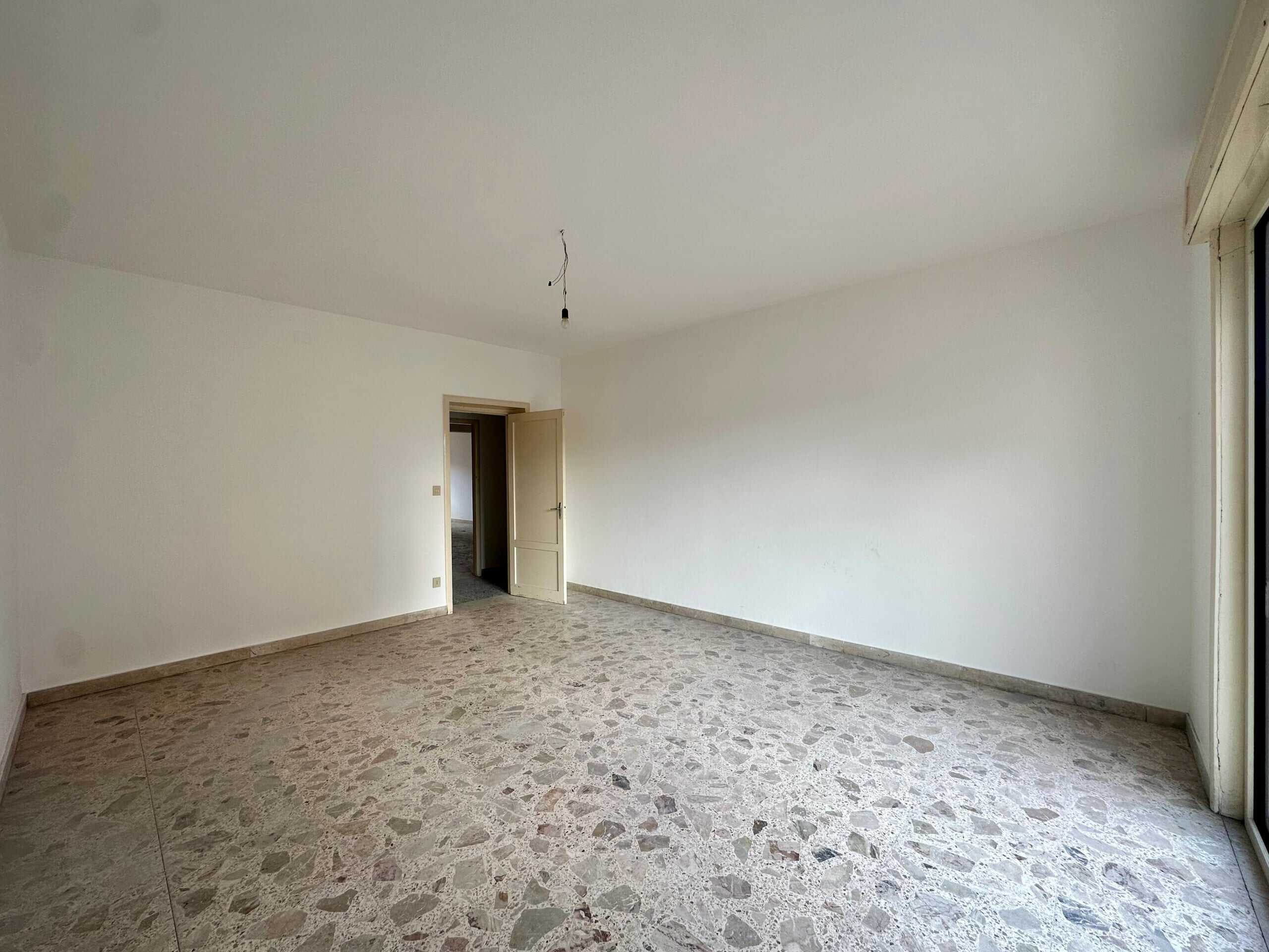 Ampio soggiorno vuoto con pavimento in terrazzo e pareti bianche, porta aperta verso un'altra stanza, grande finestra o porta scorrevole per l'accesso alla luce naturale.