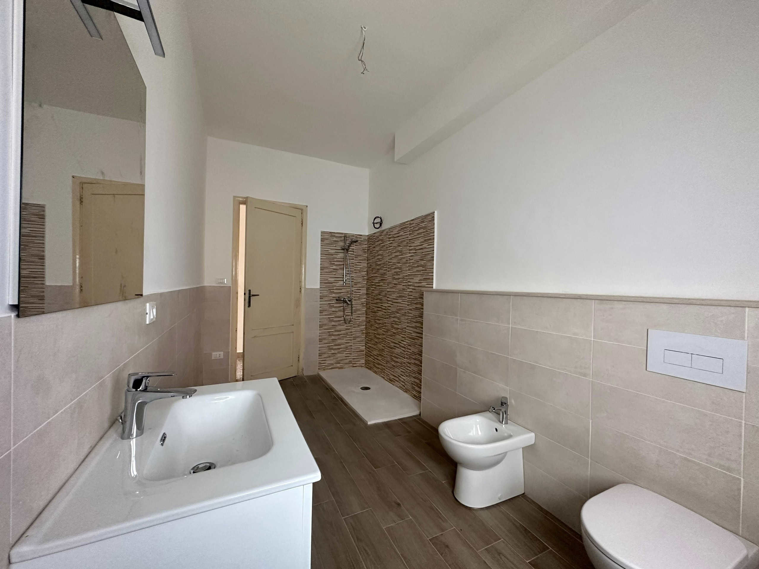 Lavanderia moderna con lavabo, bidet, wc e zona doccia in fase di completamento, con pareti rivestite in piastrelle beige e pavimento in legno effetto parquet.