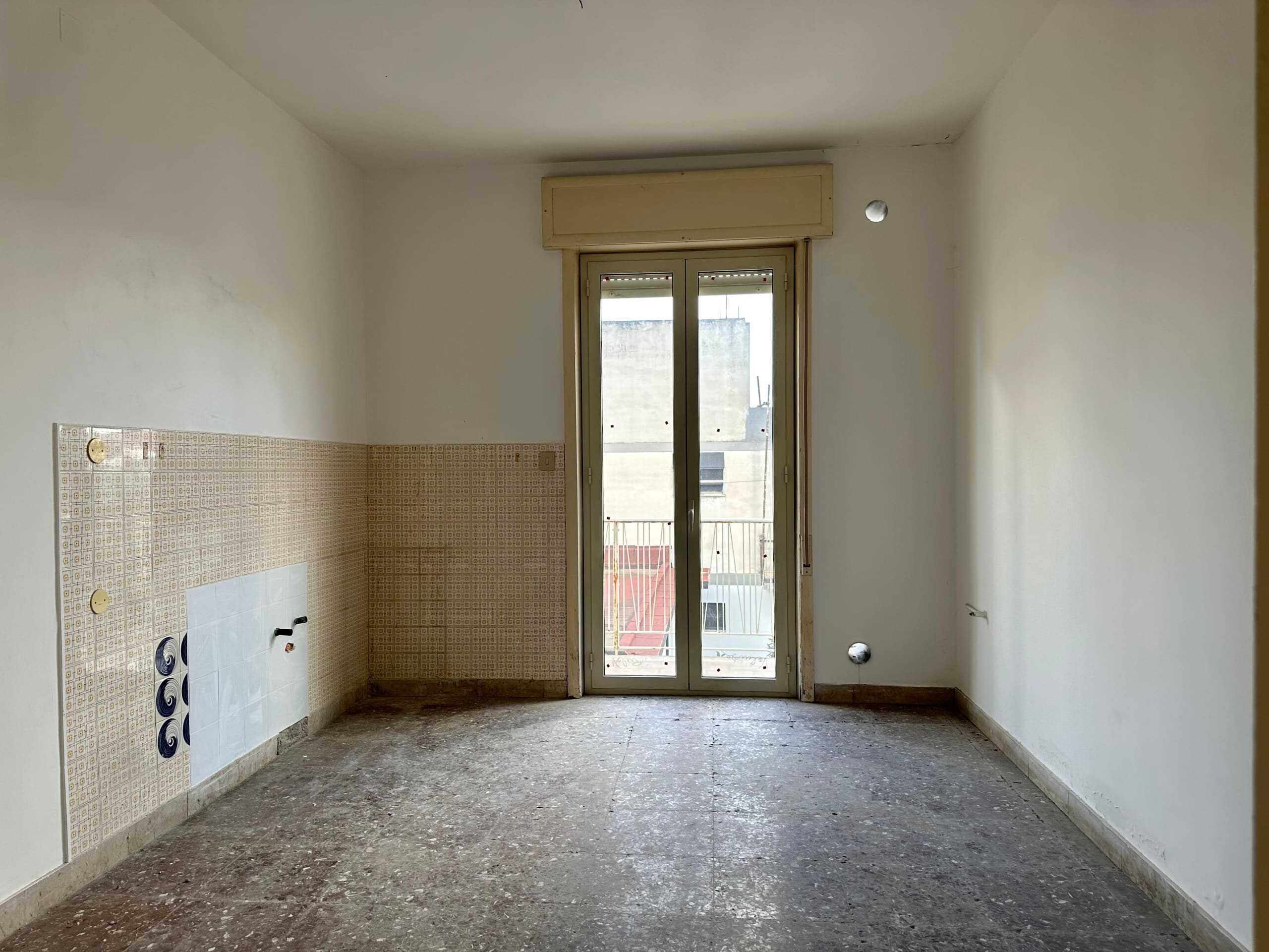 Locale vuoto con porta finestra che dà su un balcone, pareti bianche e pavimento in pietra, pronto per ristrutturazione, ideale come appartamento o investimento immobiliare.