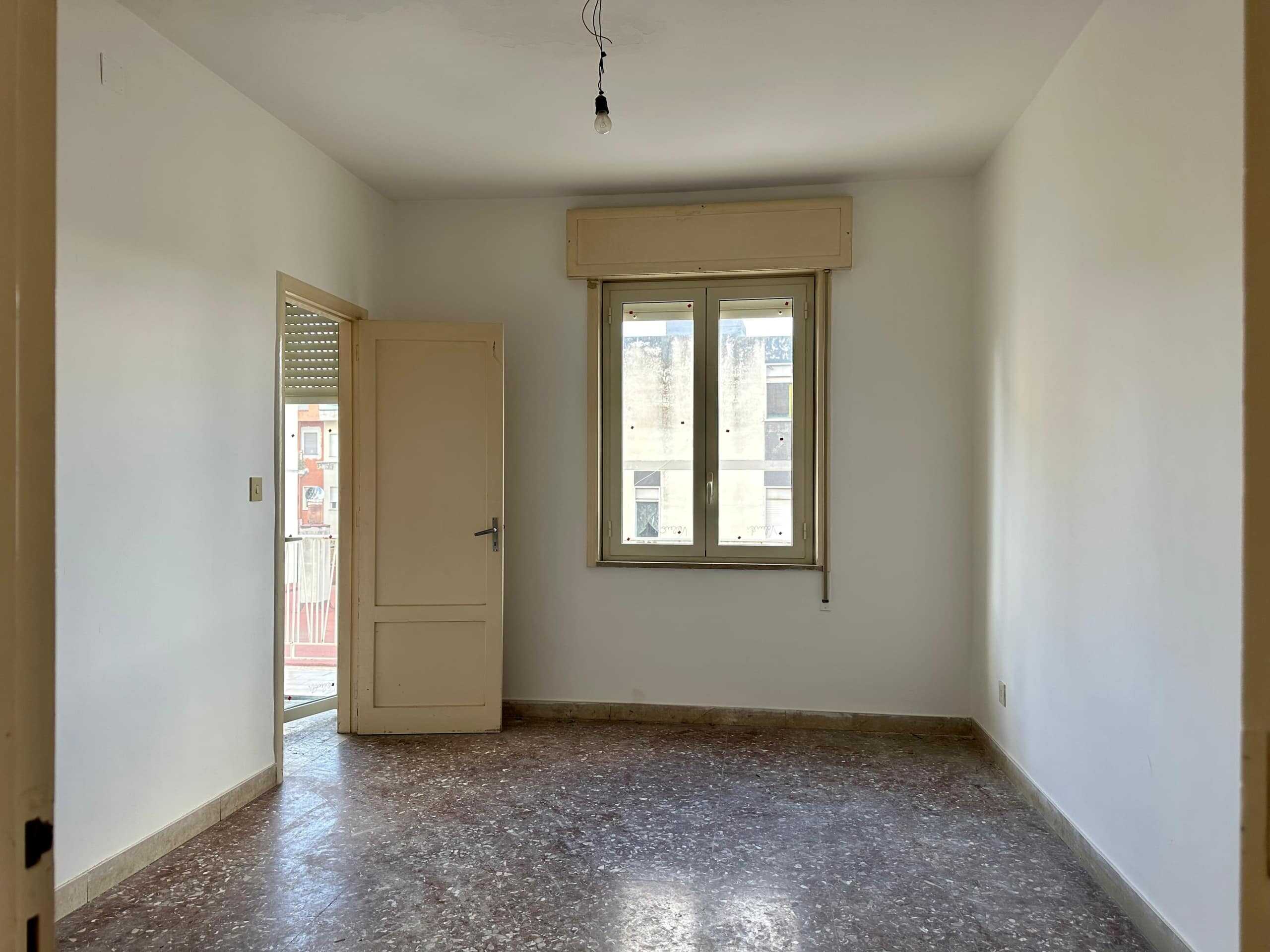 Spazio abitativo vuoto con pareti bianche, finestra e porta che conduce a un balcone, pavimento in marmo o piastrelle, ideale per ristrutturazione o arredamento moderno.