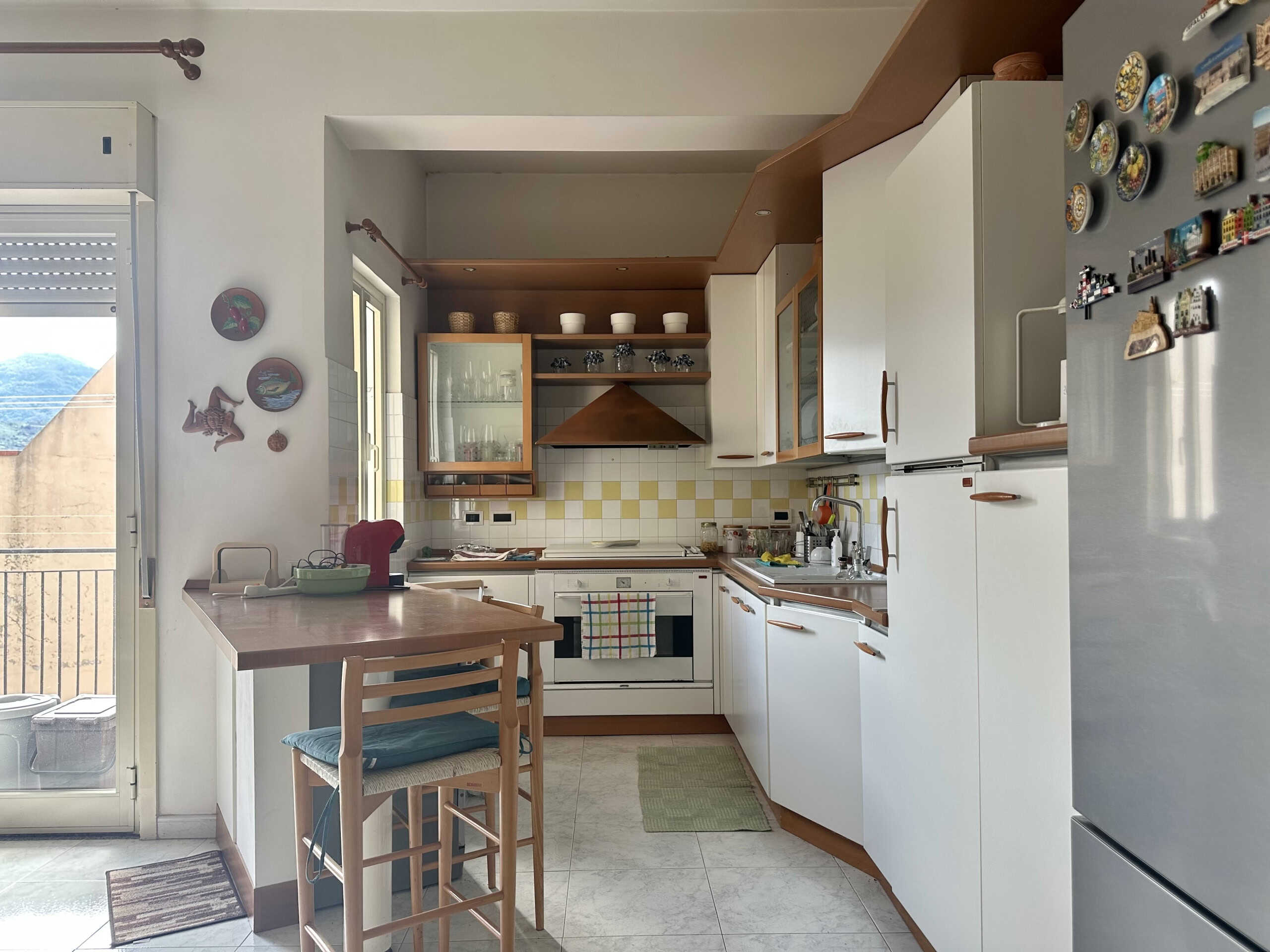 Lavagna da cucina moderna con mobili bianchi, piano di lavoro in legno, frigorifero e finestra, ideale per appartamenti e case in vendita o affitto.