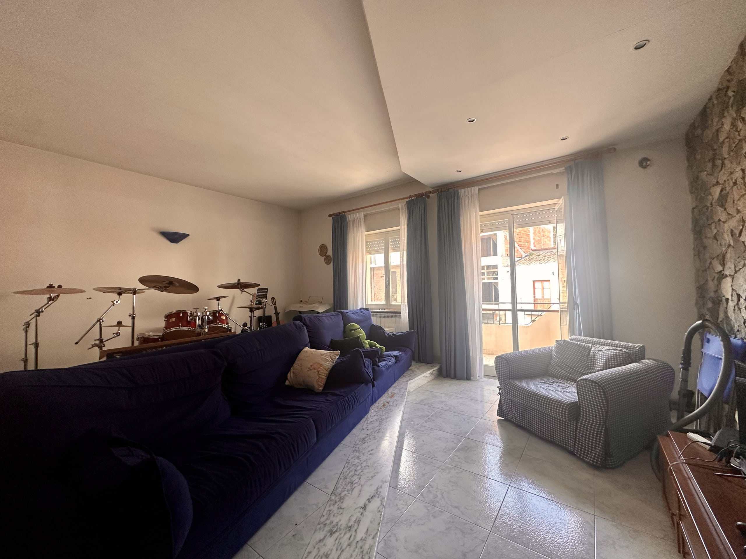 Accogliente salotto con grande divano blu, strumenti musicali, poltrona a quadretti e ampie finestrature con tende, ambiente luminoso e confortevole, ideale per momenti di relax e musica.