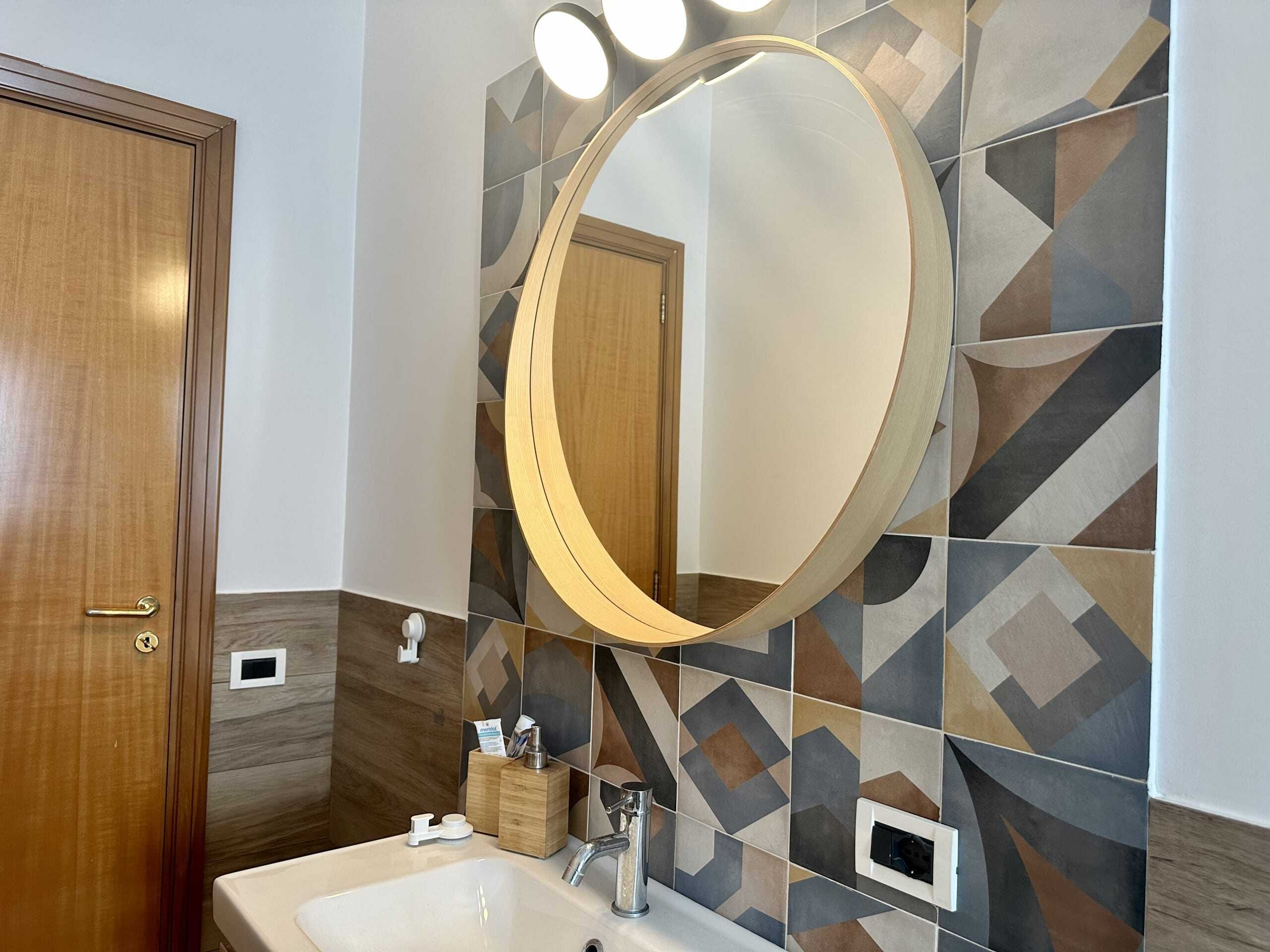 Specchio rotondo moderno in bagno con cornice in legno, moderna illuminazione a faretti e pareti decorate con piastrelle geometriche colorate, ideale per ambienti di design e stile contemporaneo.