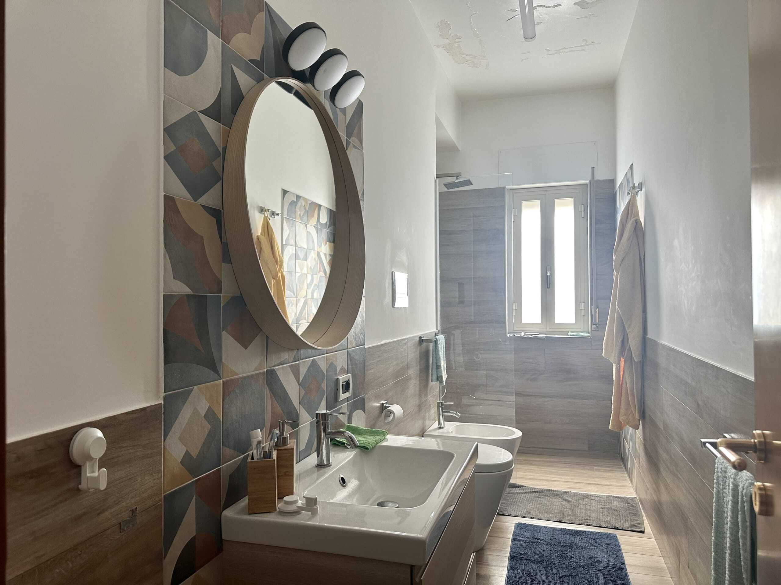 Doccia moderna in bagno con lavabo e specchio rotondo, illuminazione naturale, stile minimalista, rivestimenti in piastrelle geometriche, spazio funzionale e luminoso, ideale per edilizia residenziale e ristrutturazioni.