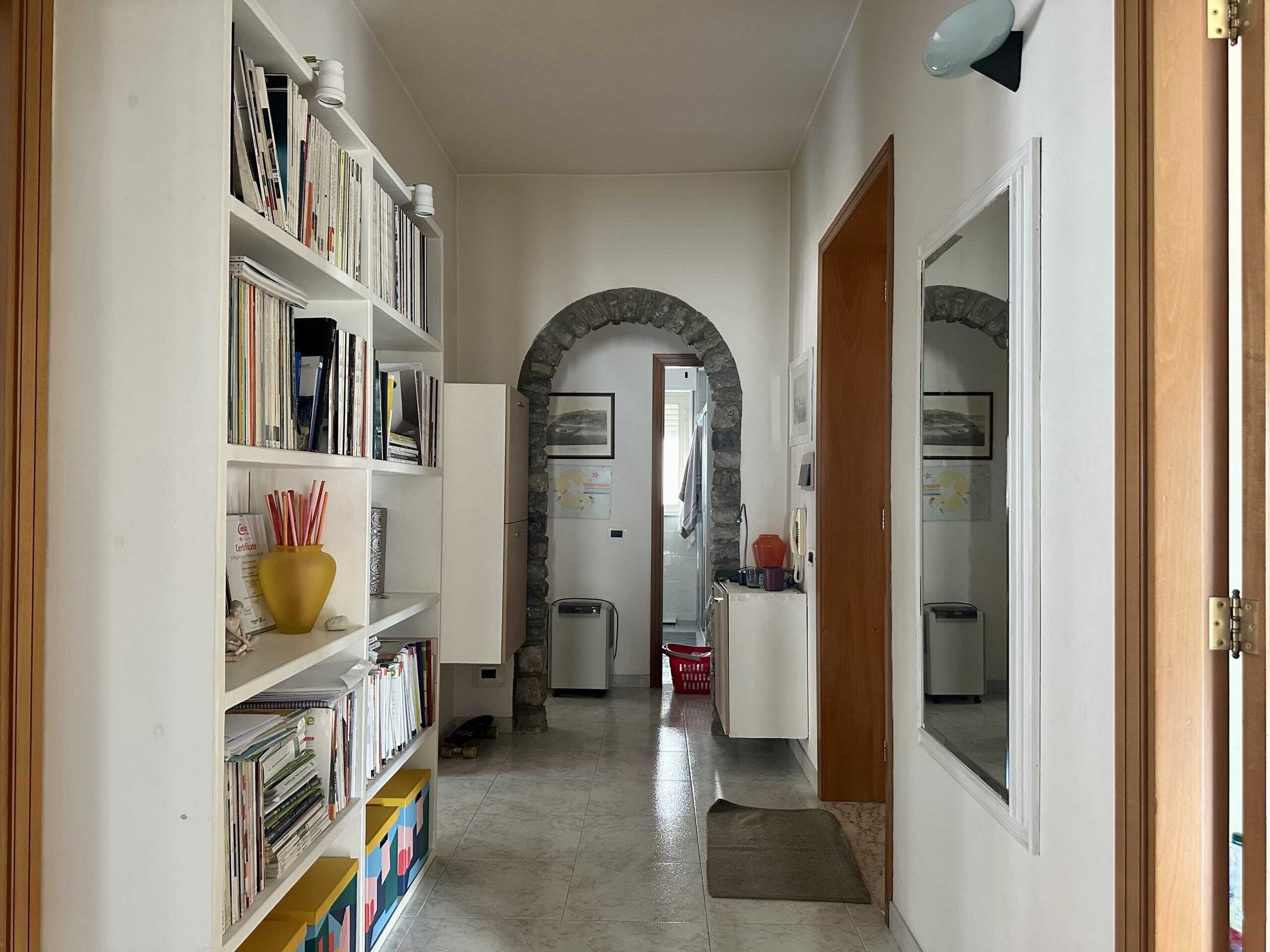 Esterno di un appartamento con porta d'ingresso, pareti bianche, libreria con libri e decorazioni, arco in pietra che conduce alla zona lavanderia, ambiente luminoso e accogliente.