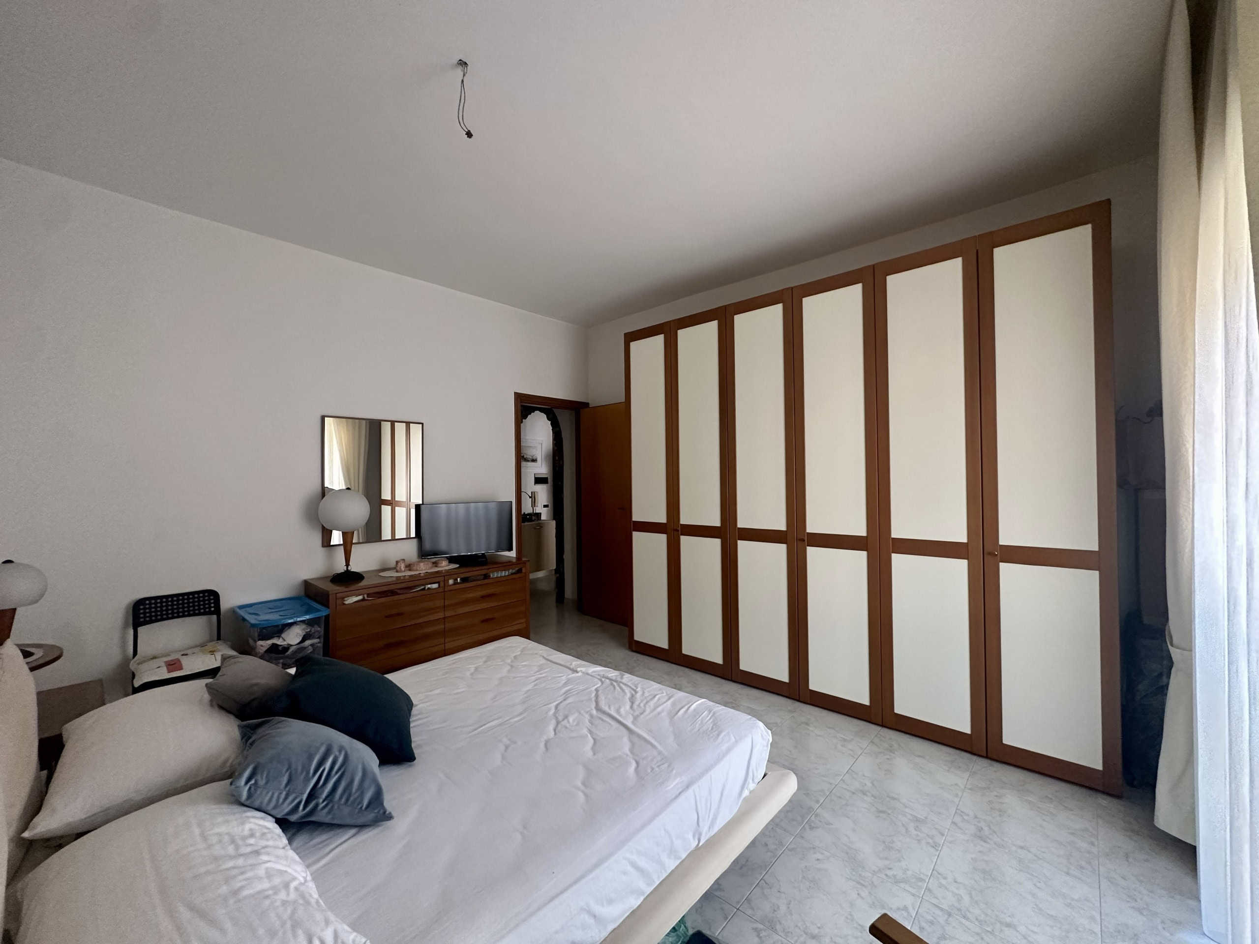 Armadio a muro in legno e bianco in camera da letto moderna con letto matrimoniale, lampade da comodino, TV e specchio. Soggiorno luminoso con accesso alla luce naturale tramite una finestra con tende.