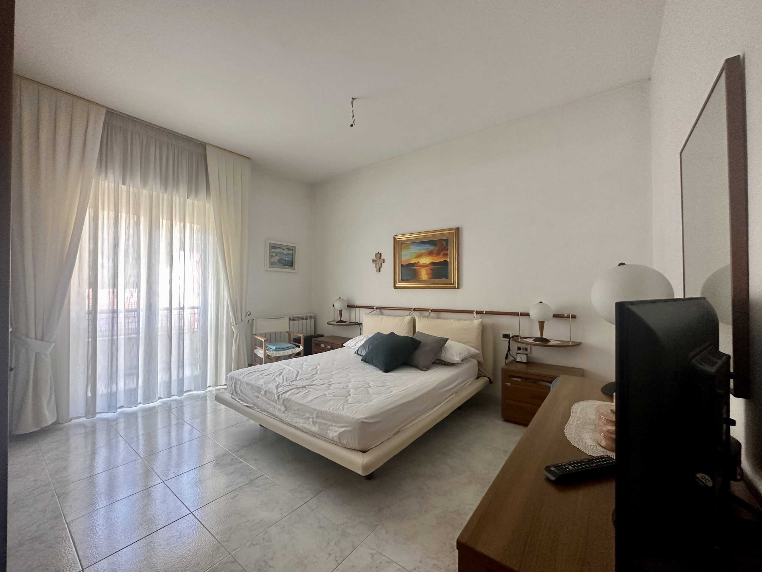 Camera da letto moderna con letto matrimoniale, tende bianche e decorazioni artistiche, ambiente luminoso e accogliente, arredamento minimalista, ideale per case e appartamenti a Roma.