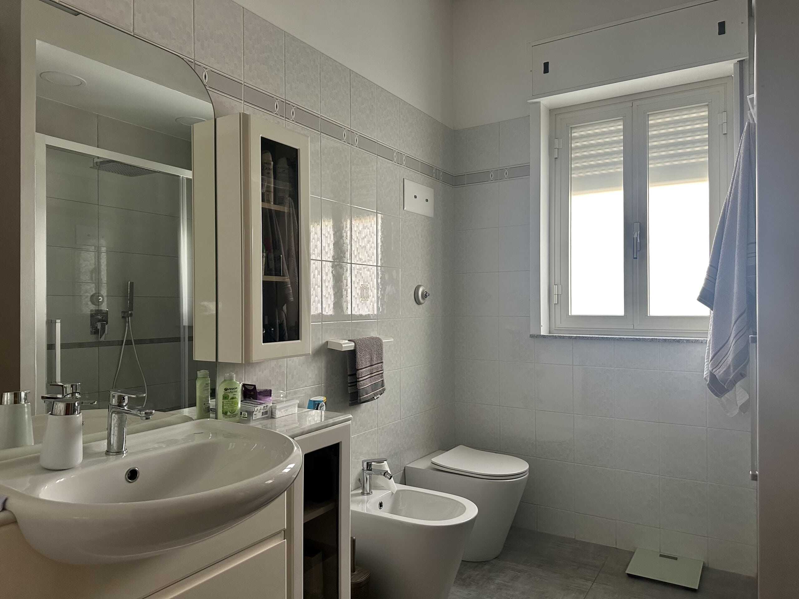 Bagno moderno con lavabo, bidet, WC e doccia, illuminato da finestra naturale, con arredi bianchi e piastrelle chiare, perfetto per ambienti residenziali di alta qualità.