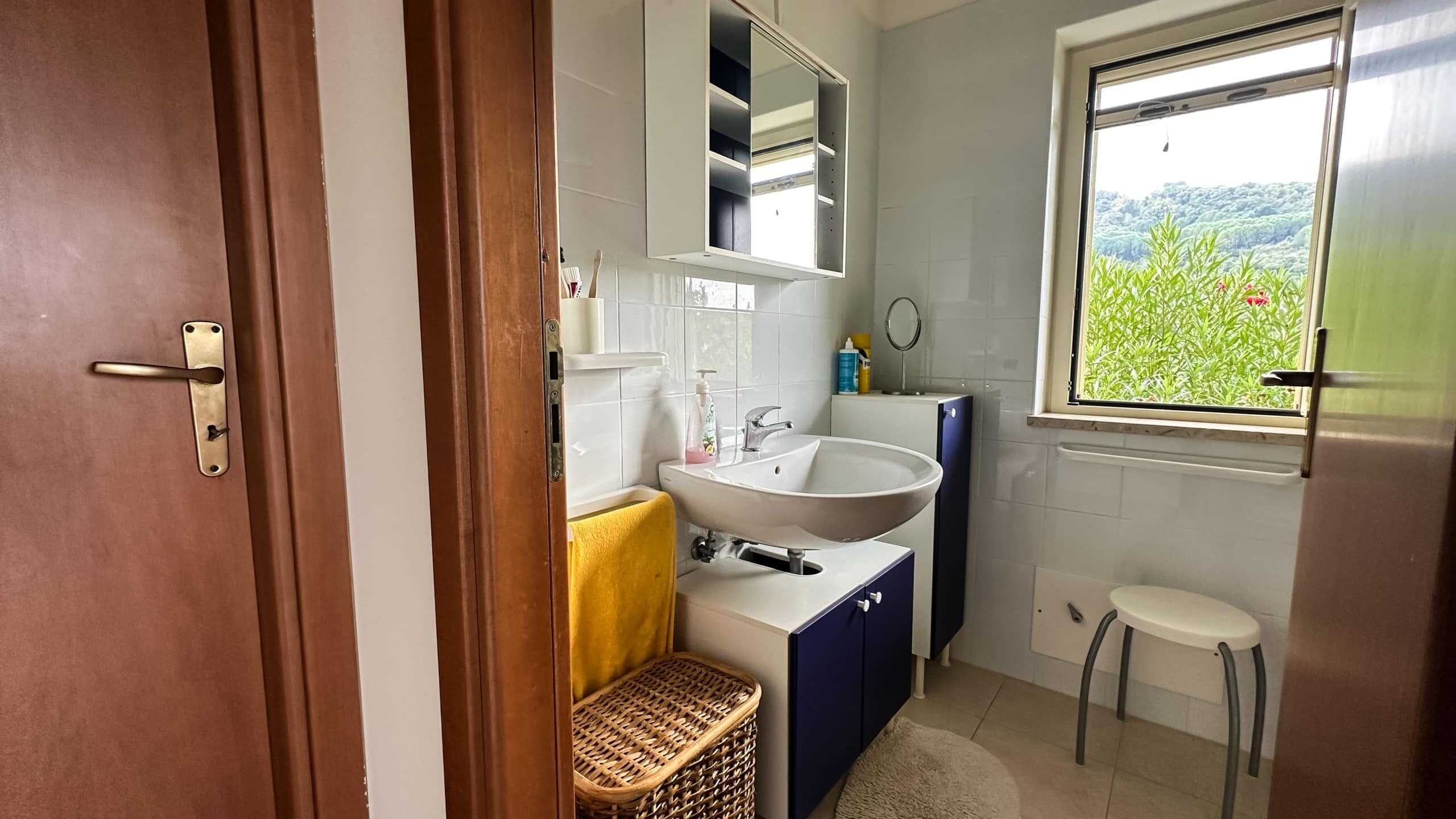 Lavabo bagno in stile moderno con mobili blu e specchiera, finestra con vista sul verde, arredo funzionale e accogliente per abitazione NextCasa.