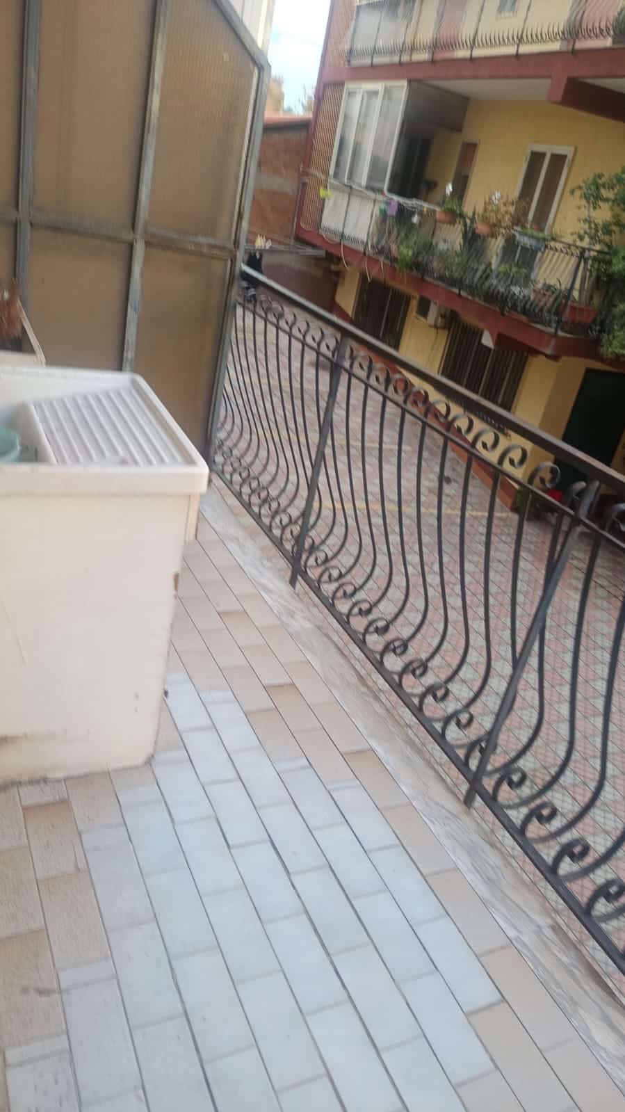 Balcone di un appartamento con grate in ferro battuto, vista su cortile condominiale con edifici residenziali, pavimentazione in piastrelle colorate, ambiente urbano, comfort e sicurezza.