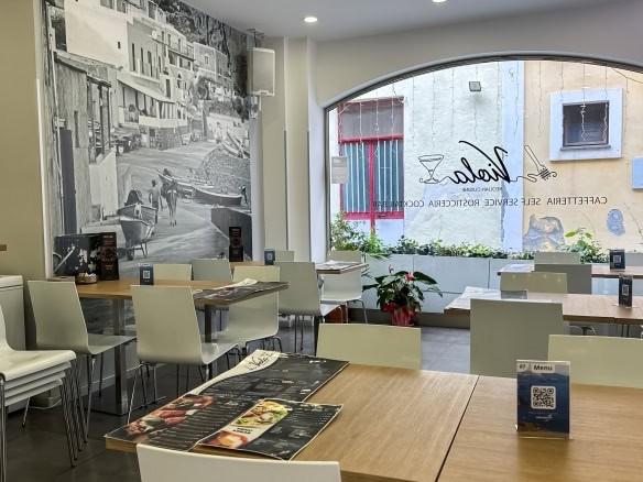 Spazio interno di un ristorante con tavoli eleganti, sedie bianche e menu, grande finestra con vista urbana, decorazioni moderne e ambiente accogliente, ideale per pranzi e cene di qualità.