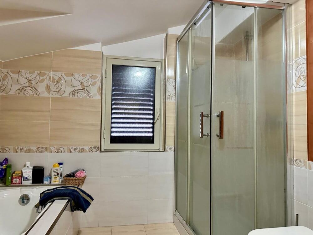 Bagno con box doccia in vetro e finestra, arredo semplice e funzionale, pareti rivestite con piastrelle beige e decorativi, ideale per ambienti di comfort e praticità, periferie residenziali.