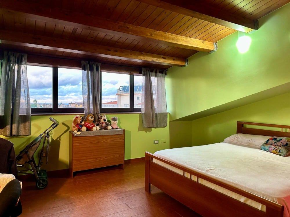 Letto matrimoniale in camera da letto con soffitto in legno e pareti verdi, spazio accogliente e luminoso, con vista città e accessori per bambini e neomamme.