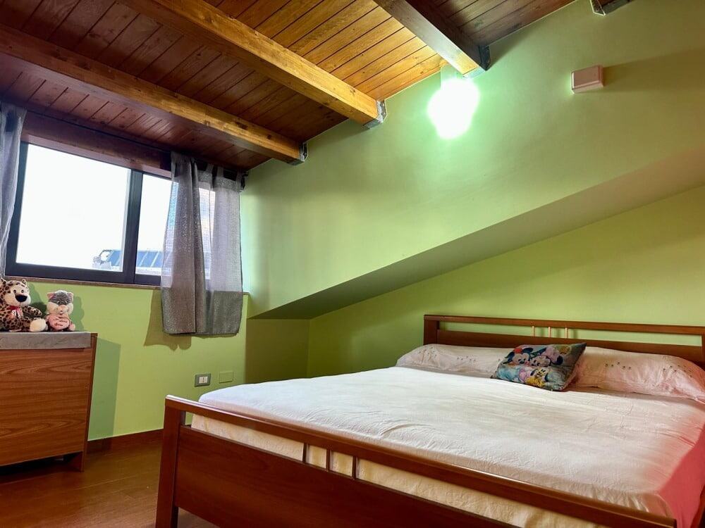 Camera da letto in mansarda con pareti verdi, letto matrimoniale, finestra con tende e soffitto in legno. Ideale per vacanze e soggiorni rilassanti, ambiente accogliente e luminoso.