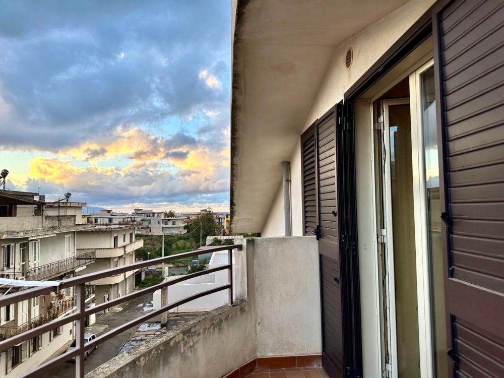 Balcone con vista urbana al tramonto, finestre con tapparelle scorrevoli, appartamento in vendita o affitto, immobile casa NextCasa, proprietà residenziale moderna, panorama città, cielo nuvoloso.
