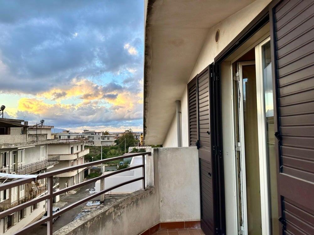 Balcone di un appartamento con vista su un complesso residenziale e un cielo nuvoloso al tramonto, caratterizzato da grate di sicurezza alle finestre e pavimento in ceramica.