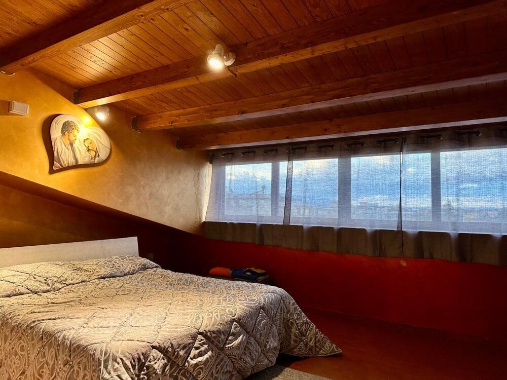 Foto di una camera da letto in stile rustico con soffitto in legno e pareti calde, grande letto con biancheria decorata e finestra con tende trasparenti che lasciano entrare la luce naturale.
