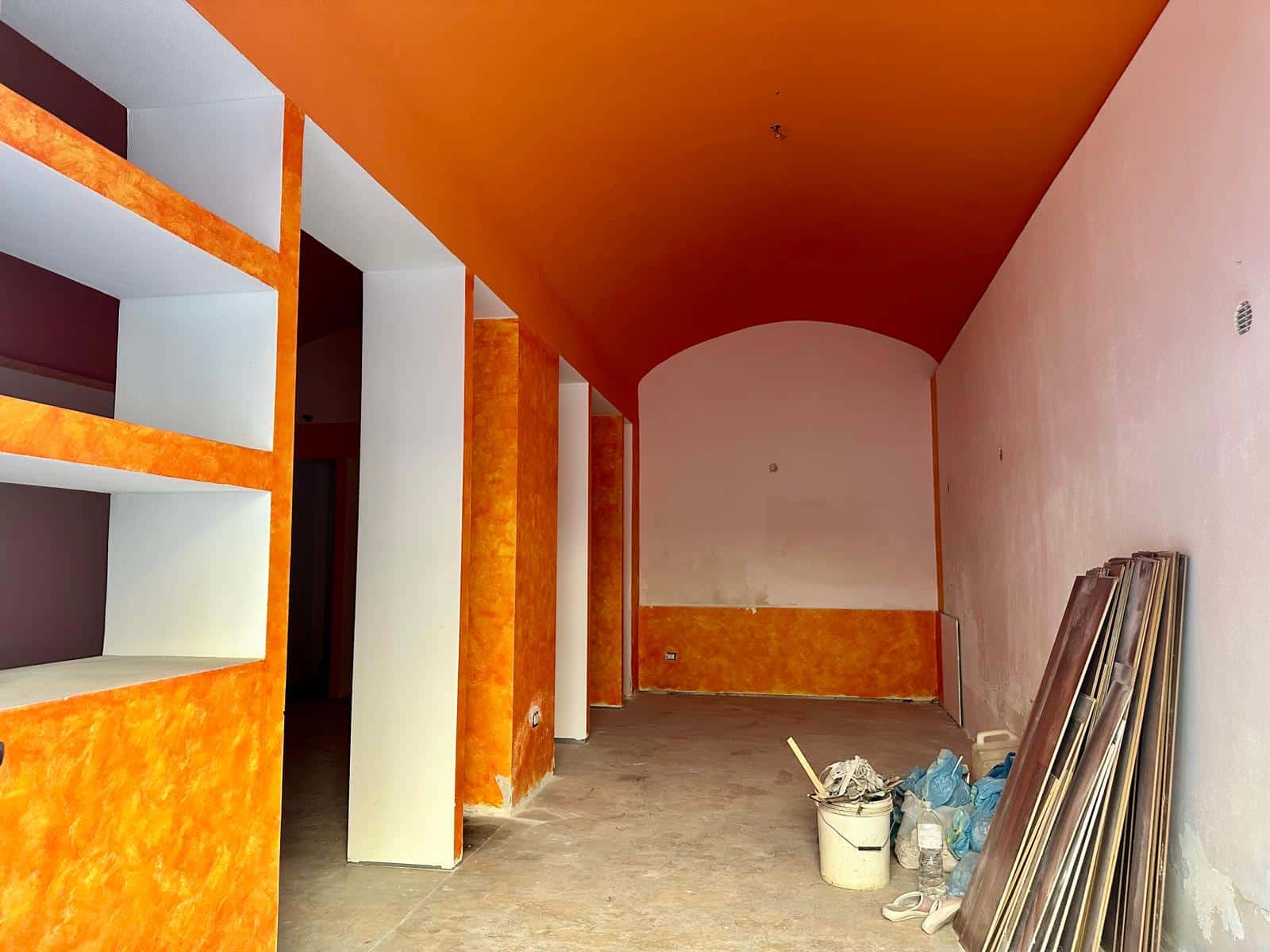 Fondo di un interno in fase di ristrutturazione, pareti e soffitto con colori arancioni e rosa, materiali da costruzione e attrezzature di cantiere appoggiate a terra, pronta per lavori di rivestimento e interior design.