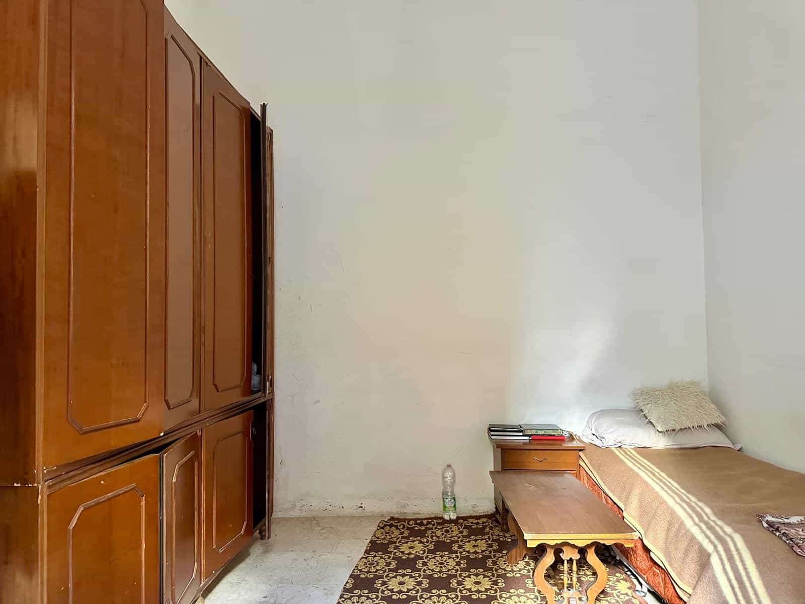 Armadio in legno massiccio con ante a battente, camera da letto semplice con letto singolo, comodino con libri e riviste, tappeto decorativo, ambiente luminoso e funzionale.