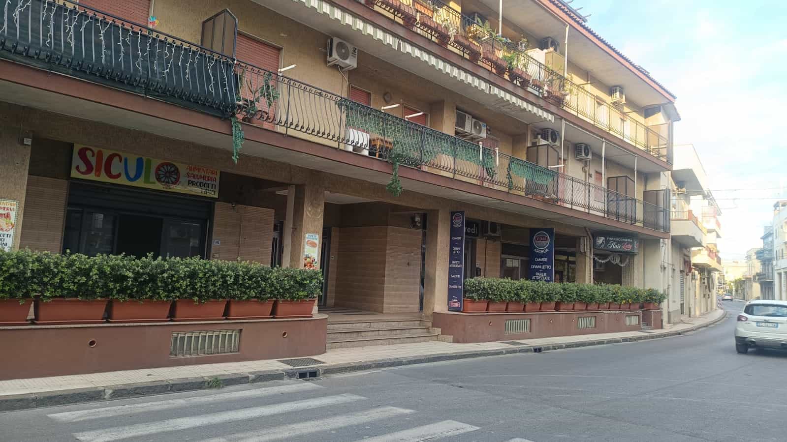 Sede di NextCasa con negozio e appartamenti in palazzo in via cittadina, piante ornamentali e balconi con piante, autoscatto lungo strada cittadina, edifici residenziali, negozio alimentare, zona urbana, architettura moderna.