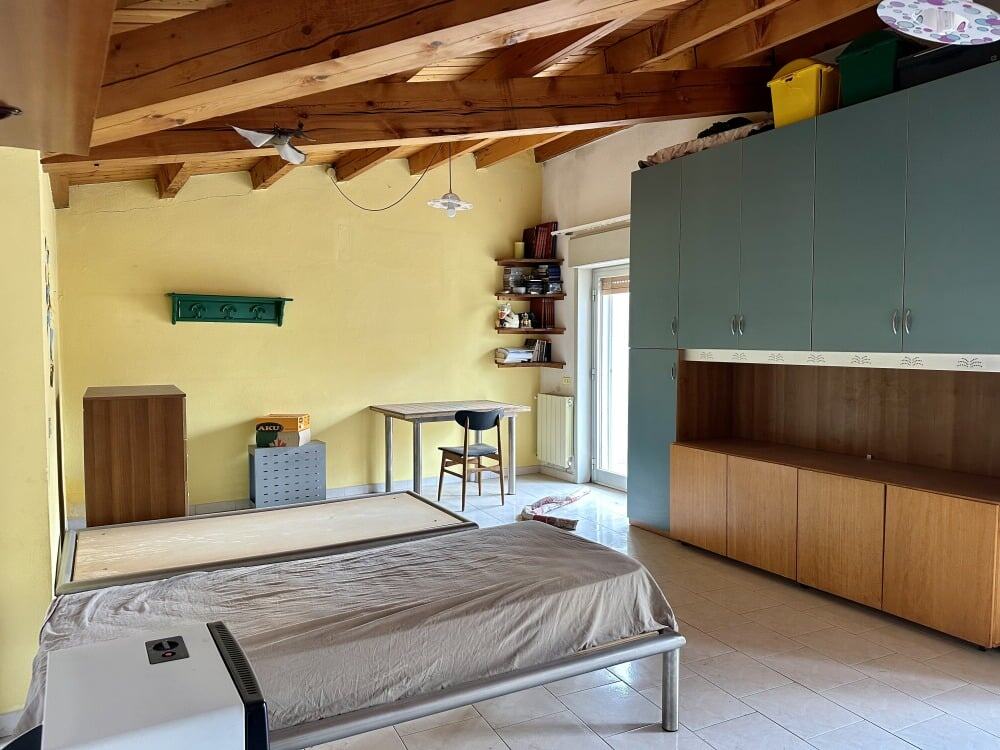 Letto singolo con materasso sfondato, pareti gialle, arredi moderni, soffitto in legno, armadio a muro con ante blu, scrivania con sedia, finestre con tende, spazio abitativo luminoso e accogliente, ideale per case in affitto o vendita.