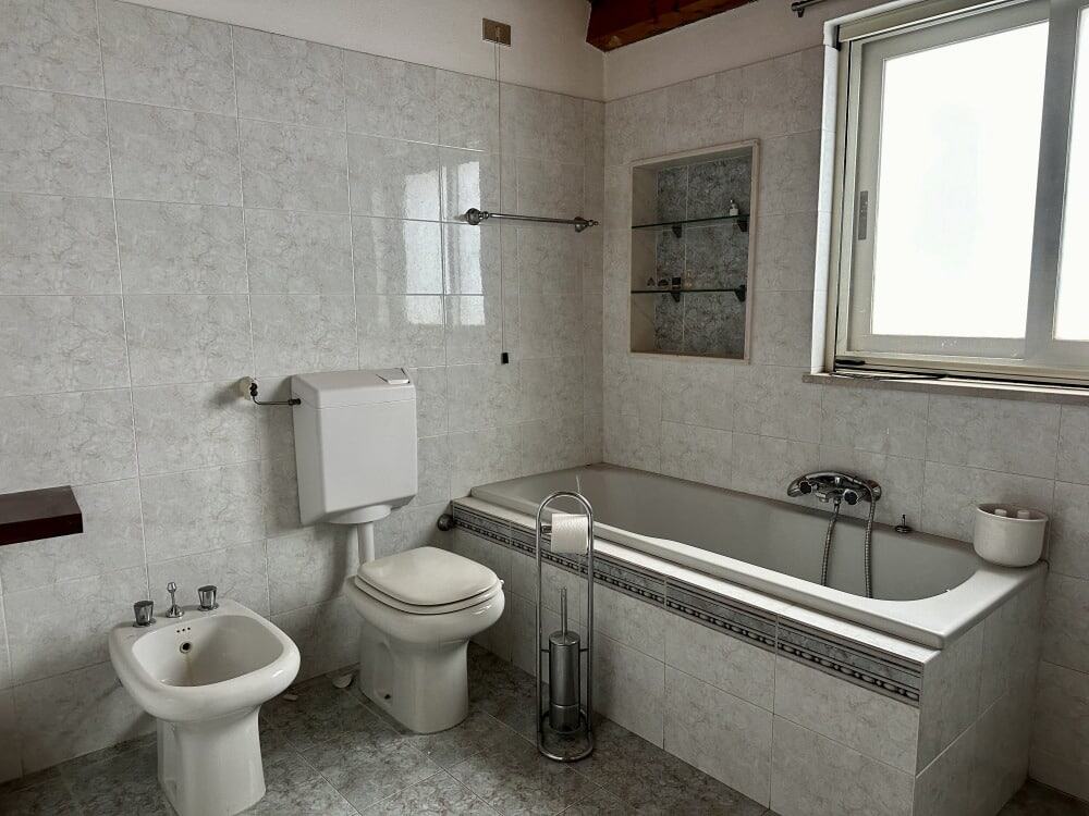 Bagno moderno con vasca, WC e bidet in arredi eleganti, pareti rivestite in ceramica chiara, finestra con infissi bianchi, zona doccia nascosta e accessori bagno minimalisti - NextCasa.