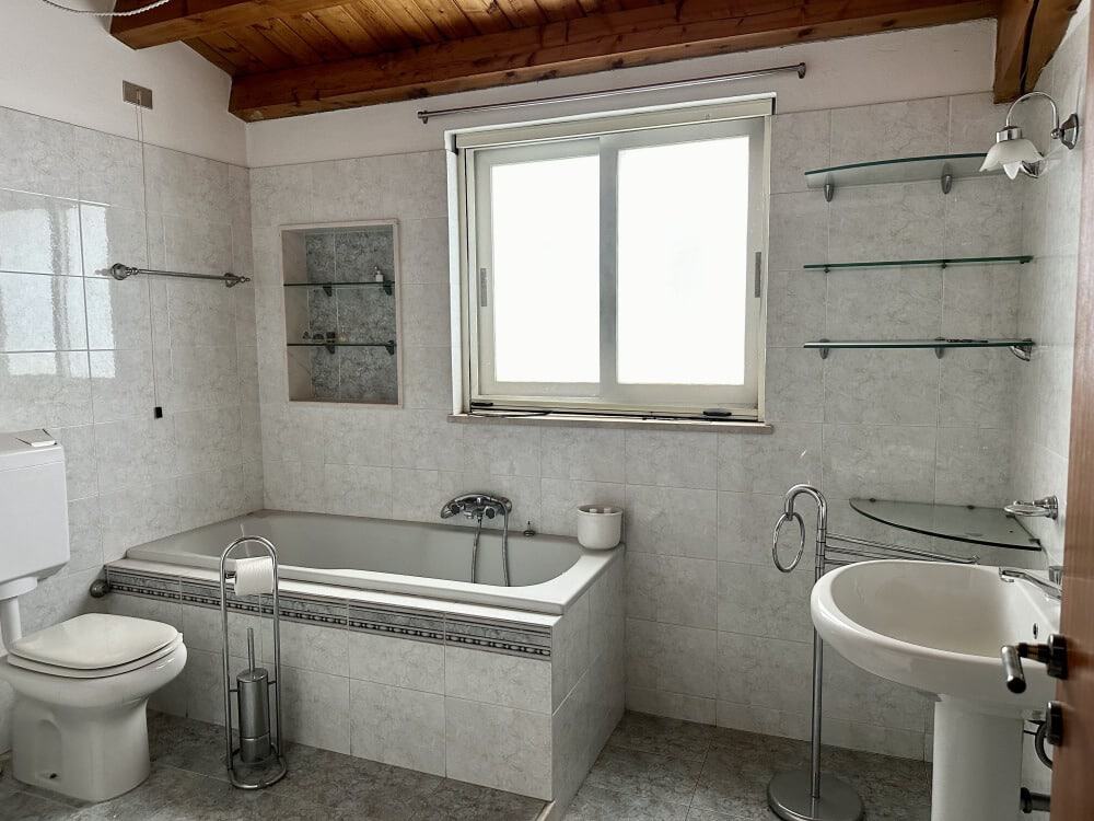 Lavabo e vasca da bagno moderna in bagno luminoso con pareti di piastrelle bianche, finestra grande, scaffali in vetro e arredi funzionali, ideale per soluzioni di arredamento bagno NextCasa.