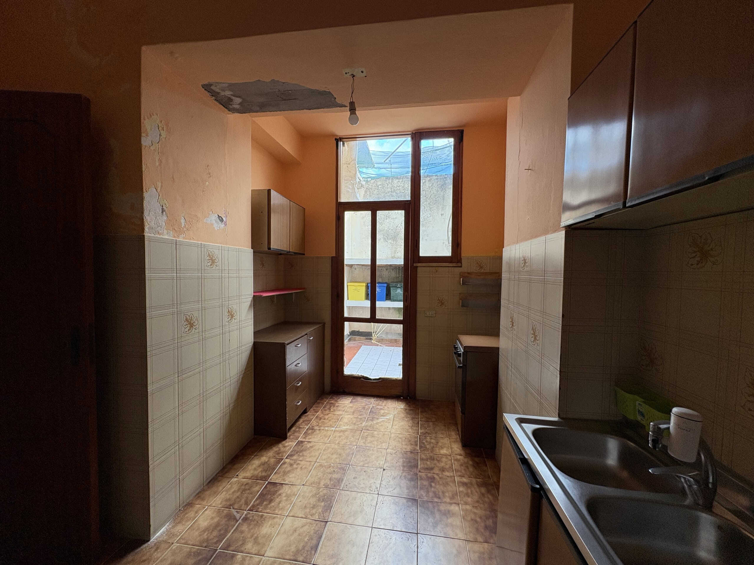 Spazio cucina in cucinino senza elettrodomestici moderni, pavimento in ceramica beige e pareti in pessimo stato, con soffitto danneggiato e finestra che apre su un cortile esterno, in un appartamento da ristrutturare.