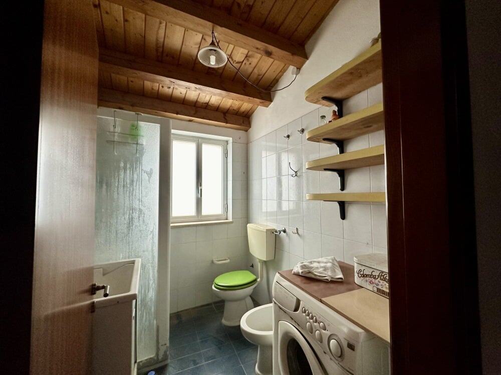 Lavanderia moderna con lavatrice, scaffali in legno, finestra panoramica e soffitto in legno, ideale per ristrutturazioni e arredo bagno, ambiente funzionale e accogliente.