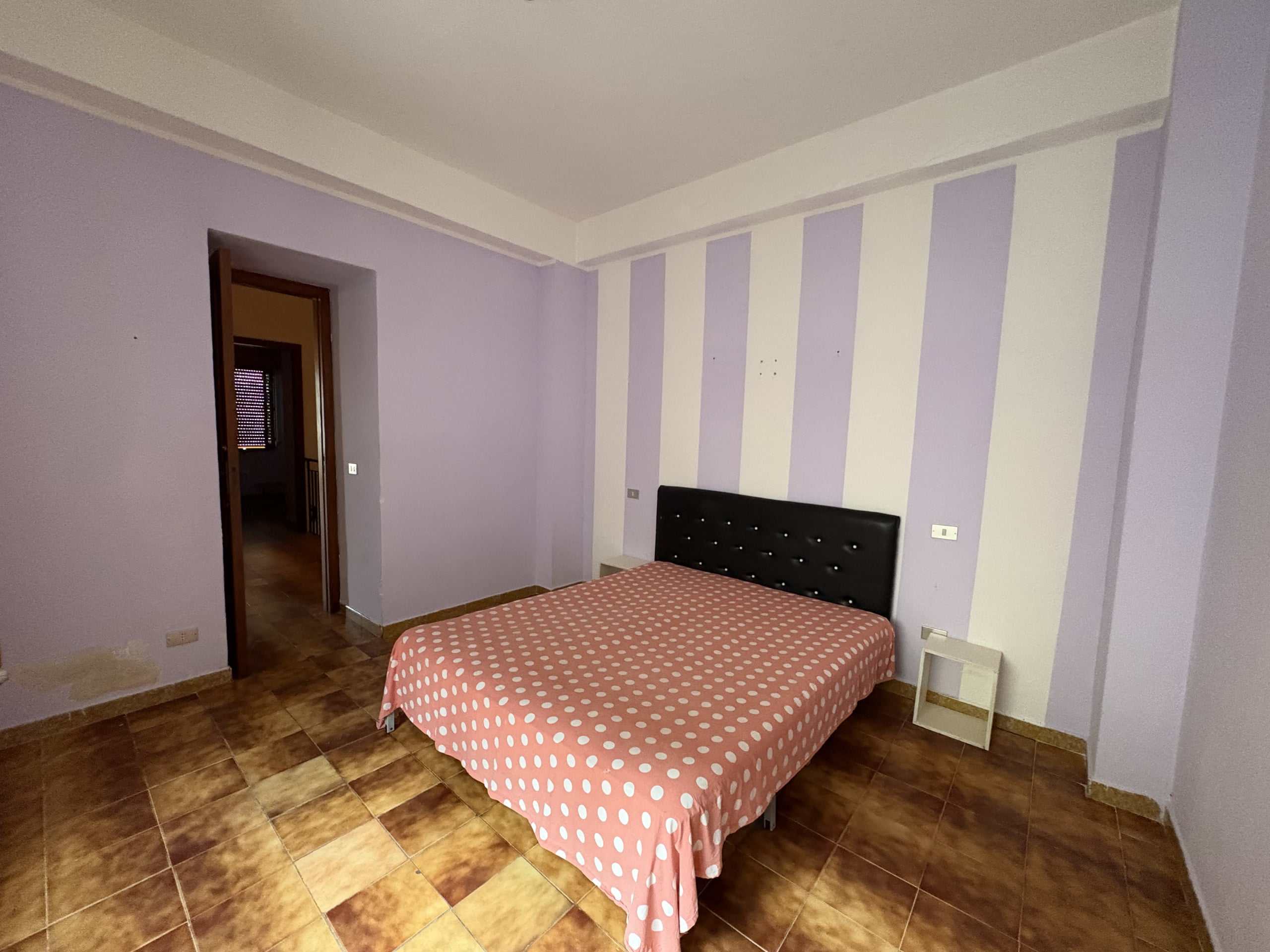 Letto matrimoniale in camera da letto con pareti viola e pavimenti in gres porcellanato effetto legno, ideale per appartamenti e case in vendita o affitto a NextCasa.