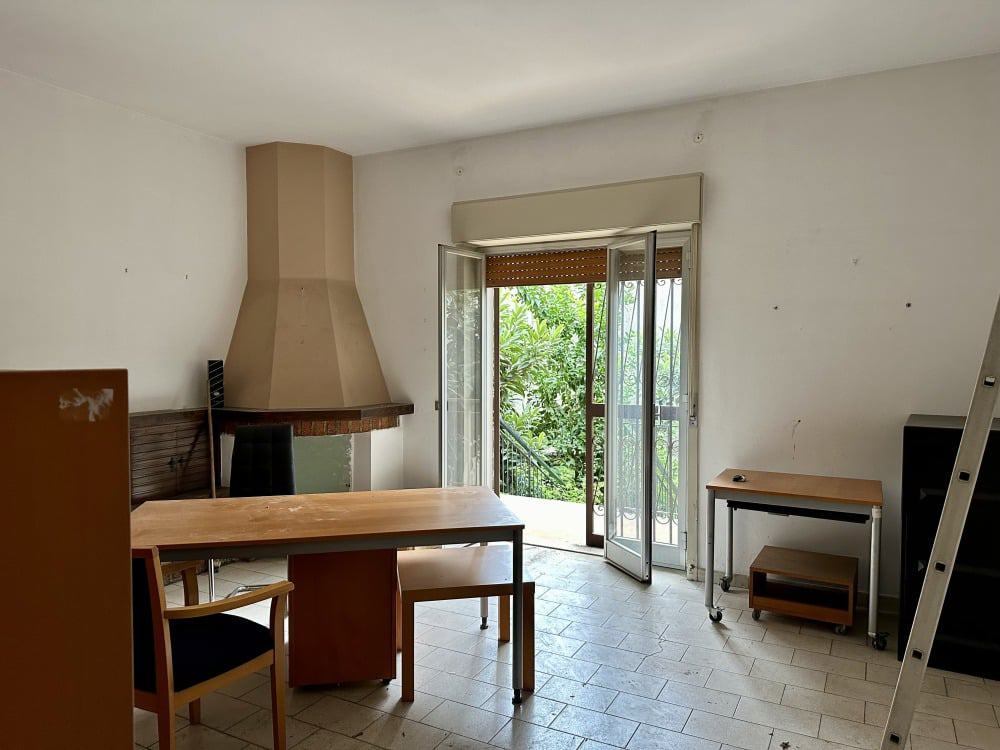 Spazio soggiorno con camino, grande finestra e balcone con vista verde, arredamento minimalista, ambiente luminoso e accogliente, ideale per abitazioni in vendita o affitto.