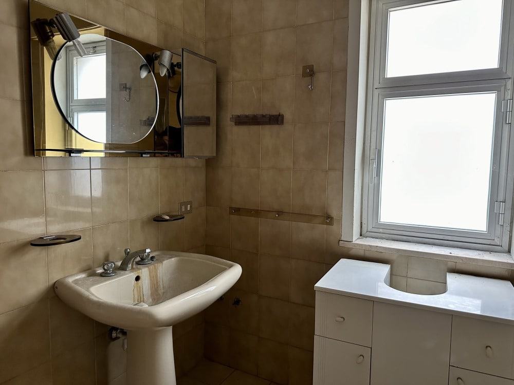 Lavabo e specchio in bagno con pareti rivestite in piastrelle beige, finestra grande, mobili da bagno bianchi, spazio funzionale e luminoso, ideale per ristrutturazioni e arredo bagno moderno.