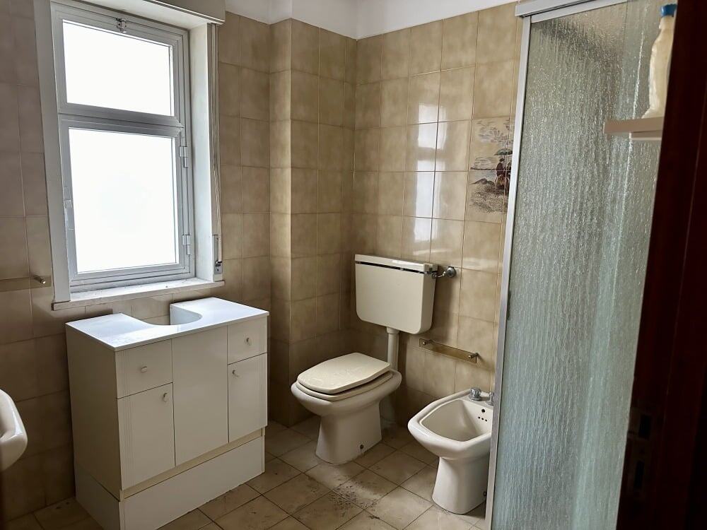 Lavabo, WC e bidet in bagno con finestra e piastrelle beige, arredo semplice e funzionale, ambiente luminoso e pratico.
