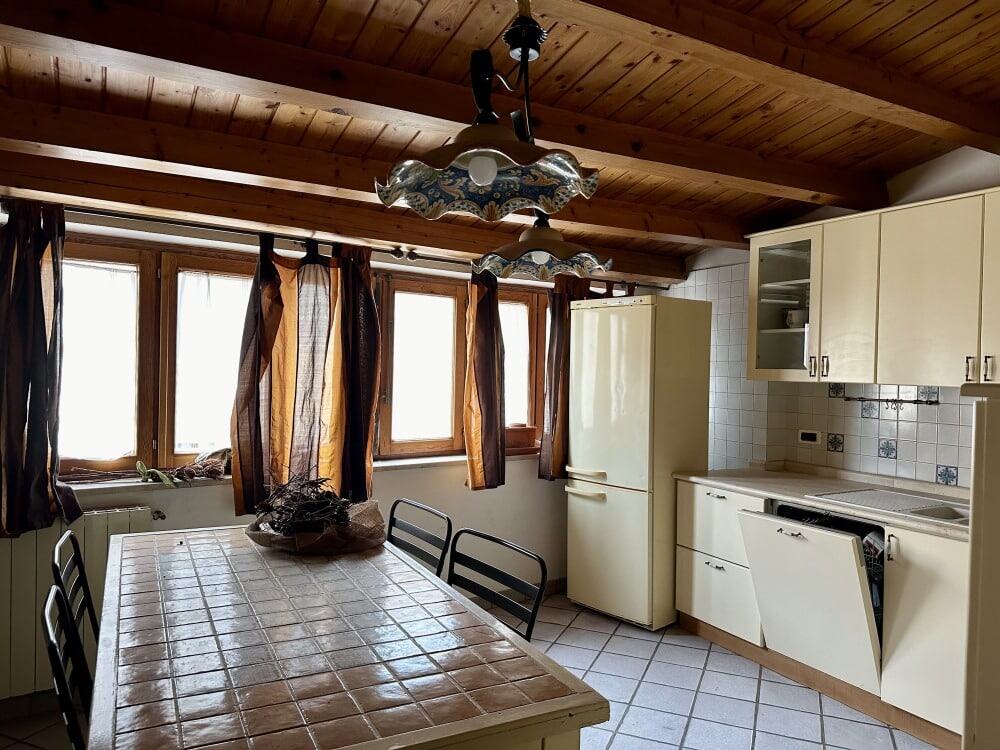 Luminoso soggiorno con grandi finestre e tendaggi castani, cucina moderna con mobili bianchi, pavimento in piastrelle e soffitto in legno, ideale per ambienti abitativi accoglienti e funzionali.