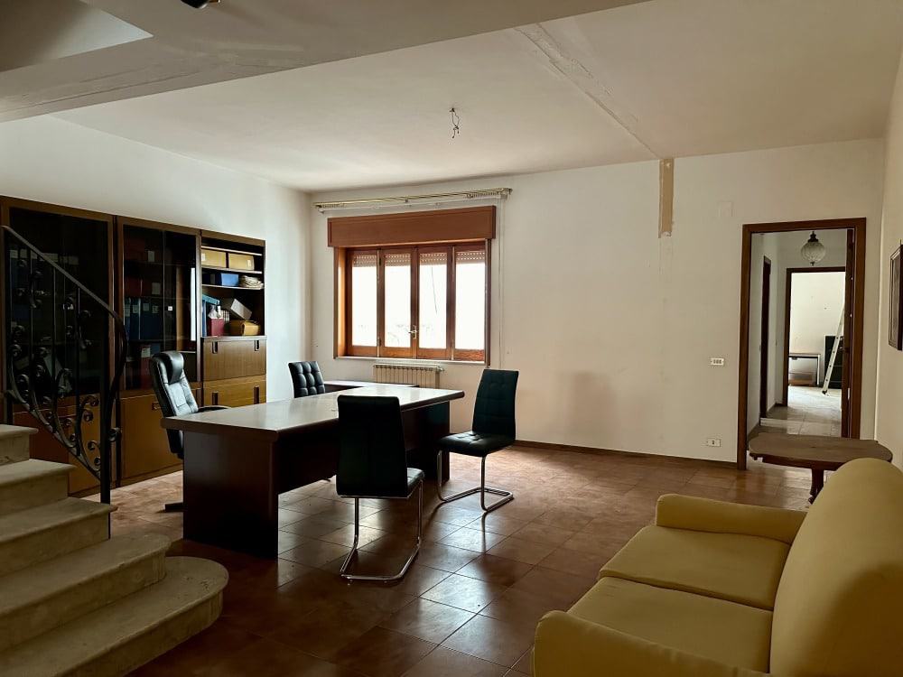 Spazio luminoso con grandi finestre, area soggiorno con divano giallo e sala da pranzo con tavolo e sedie nere, arredi classici, pareti bianche, atmosfera accogliente e funzionale.