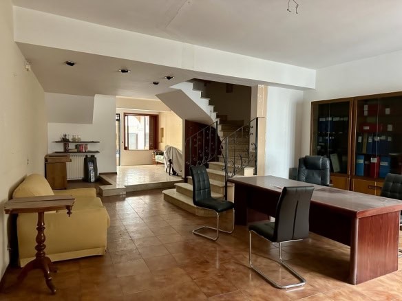 Ampia stanza con tavolo da lavoro, poltrone nere, libreria con documenti, divano beige, scala in metallo, ambiente luminoso e accogliente, arredamento funzionale e pratico, ideale per ufficio o studio domestico.
