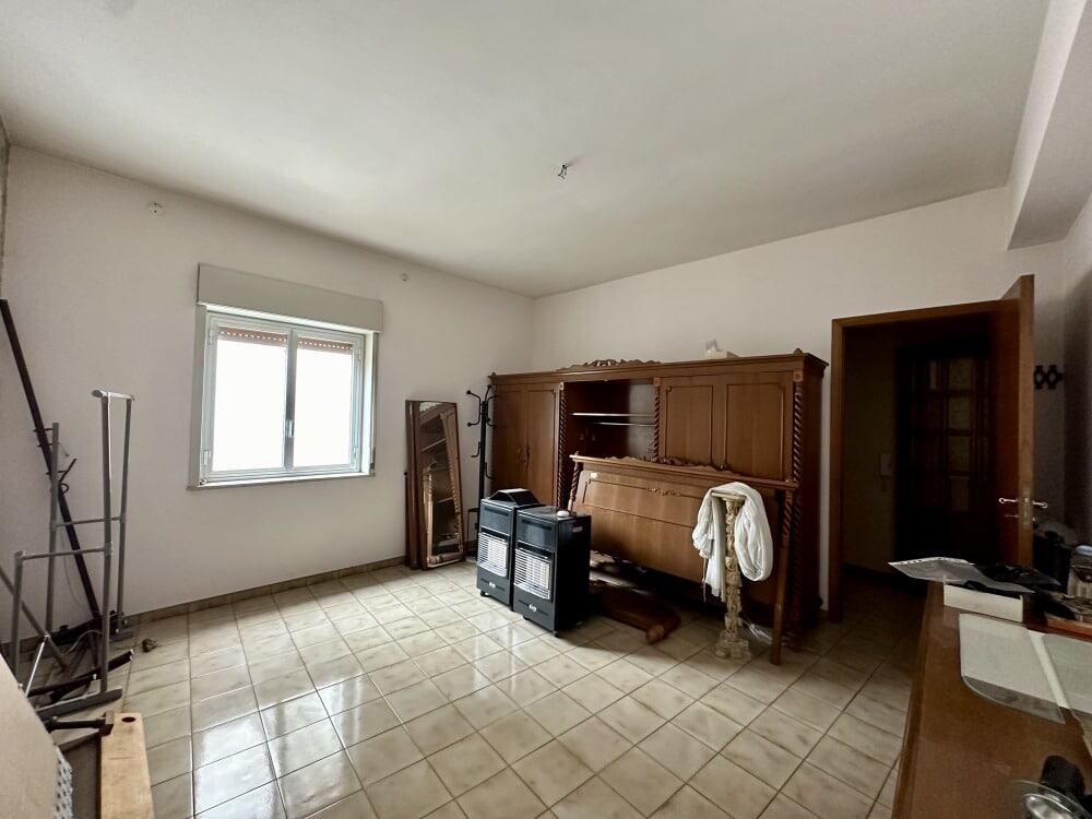 Vecchio soggiorno con armadio in legno e termosifoni, stanza di casa in fase di ristrutturazione, con piastrelle beige, finestra luminosa e porta in legno.