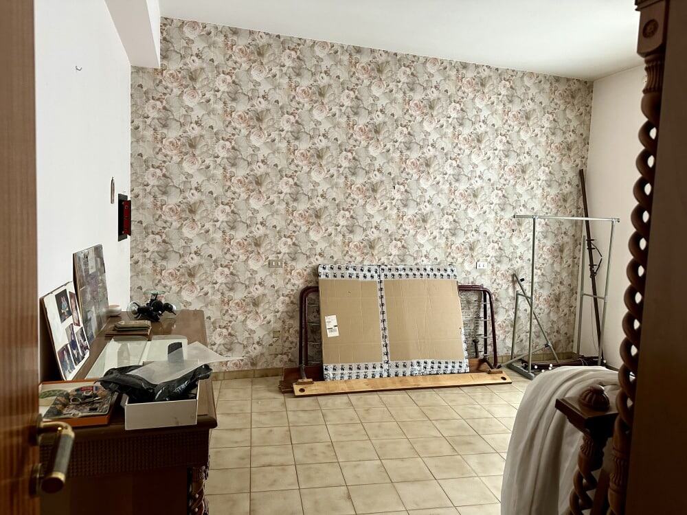 Letto e vari arredi in una stanza con parete rivestita in carta da parati floreale, piano in ceramica, e alcuni mobili e oggetti da sistemare o riparare, in ambientazione di interni abitativi.