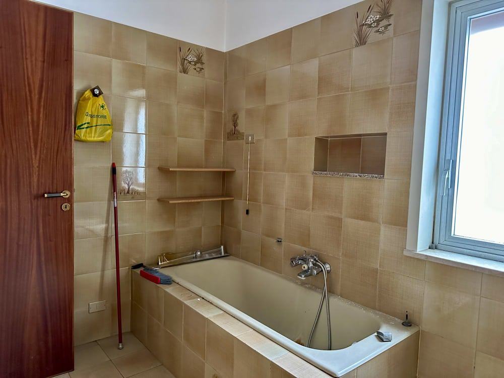 Bagno in stile classico con vasca da bagno, pareti rivestite in ceramica beige, mensole in legno, finestra grande per luce naturale, accessori per pulizia, ambiente funzionale e semplice.