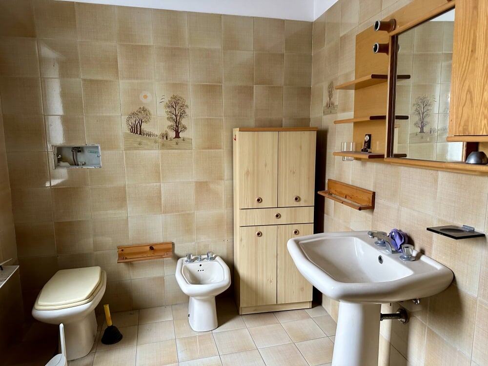 Bagno con piastrelle beige, lavabo, bidet, toilette, mobili in legno, specchio, mensole e accessori decorativi in stile semplice e funzionale.
