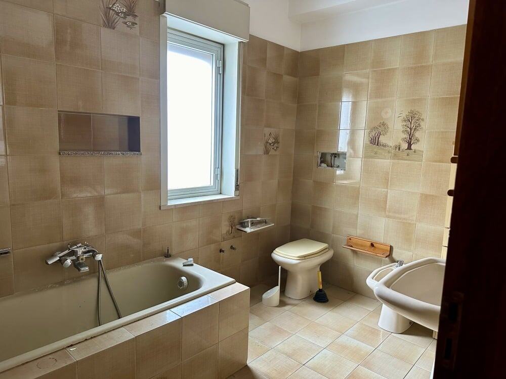 Bagno in stile classico con vasca, WC e bidet, pareti e pavimenti in piastrelle beige, finestra per aria fresca, accessori bagno essenziali, spazio funzionale e luminoso, ideale per l'arredamento e ristrutturazione.