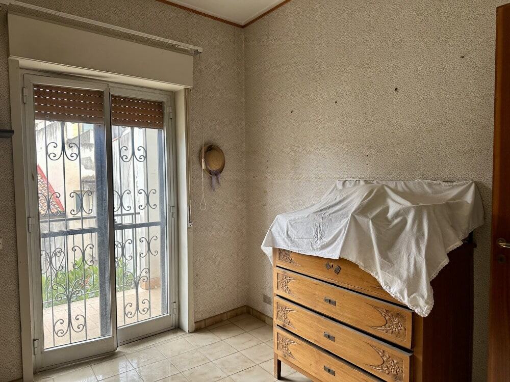 Luminosa stanza con porta finestra e ringhiera in ferro battuto, mobili in legno e pareti colorate, ideale per ambienti residenziali o investimenti immobiliari.