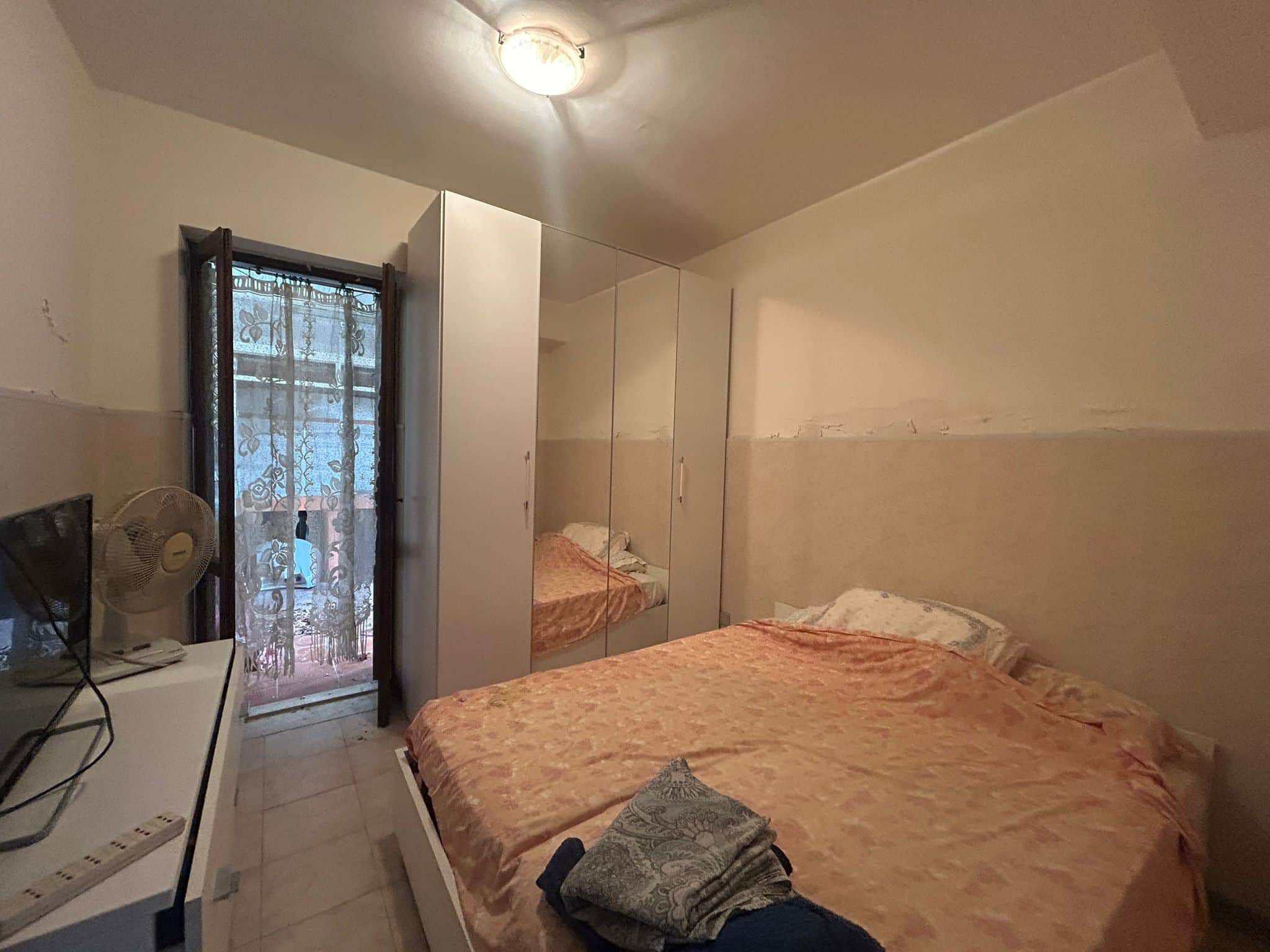 Camera da letto doppia con armadio a muro, balcone con tende floreali, letto con lenzuola arancioni, ambiente compatto e funzionale, ideale per affitto o acquisto immobiliare.