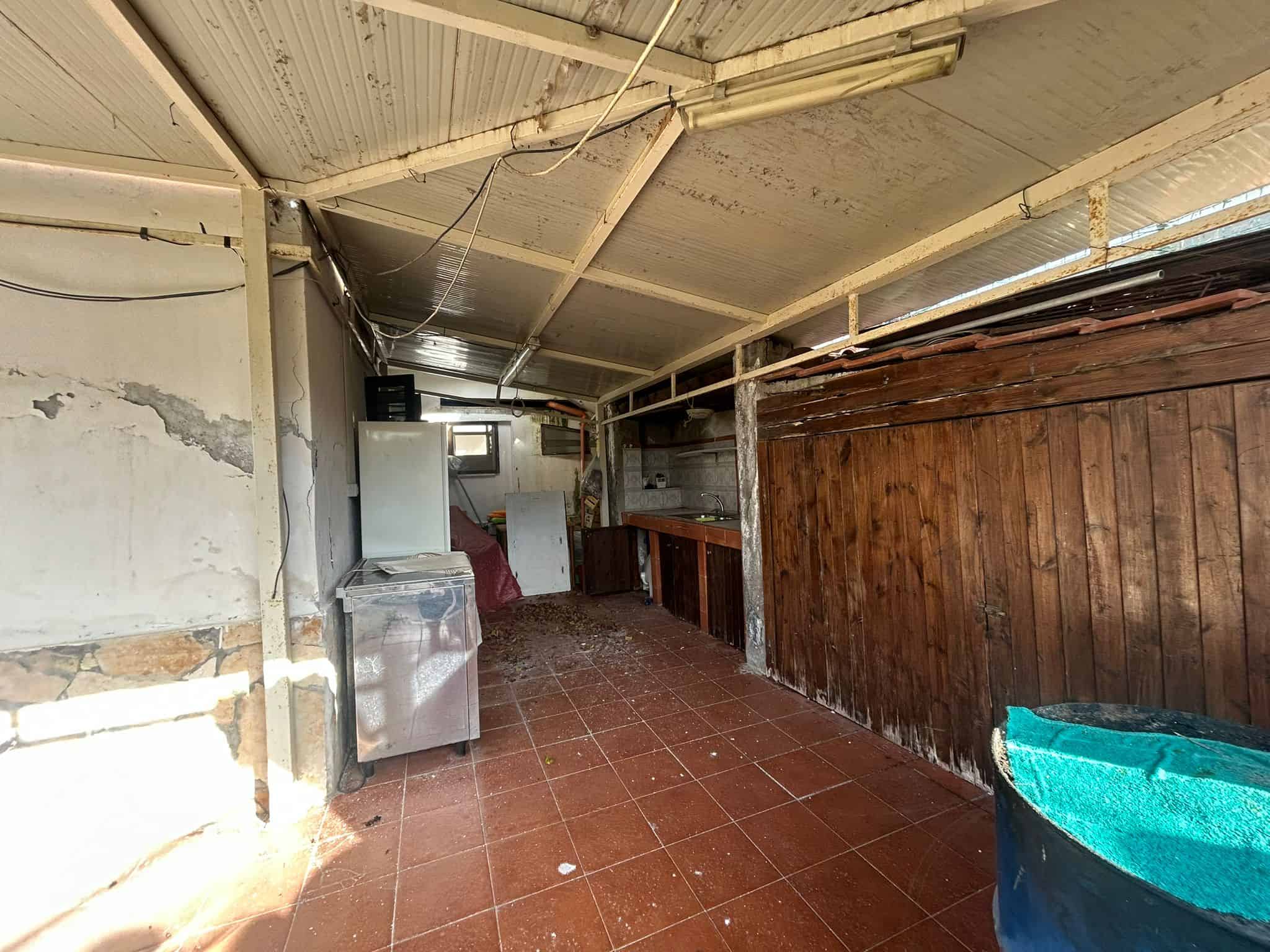 Vecchia rimessa esterna con pareti in pietra e legno, tetto in lamiera, pavimento in ceramica rossa, cucina con mobile in legno e lavello in acciaio, area di deposito o lavoro, ambientazione rustica per immobili NextCasa.